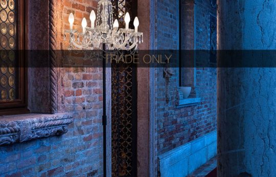 A decorative crystal chandelier hangs on a brick wall, emitting a warm glow. The setting includes a vintage-style window with intricate ironwork, along an elegantly lit corridor with textured brick walls. A banner overlay reads "TRADE ONLY.