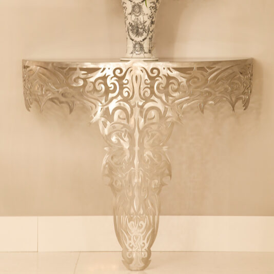 The Exclusive Limited Edition Designer Console, a decorative metal console table attached to a wall, features intricate floral and scrollwork patterns. This half-moon shaped table cascades down to the floor with a single ornate leg and holds a beautifully designed porcelain vase in blue and white.