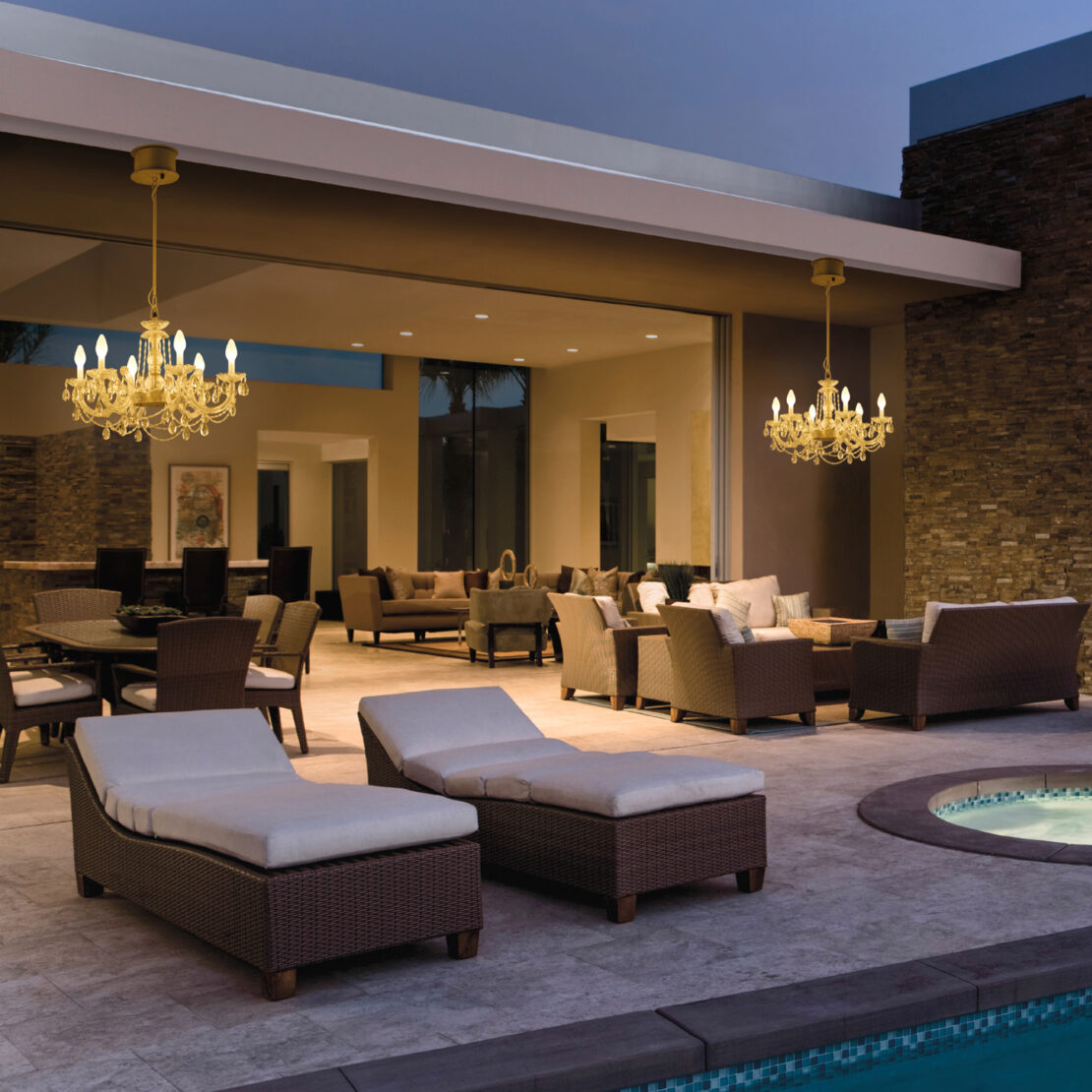 At dusk on a spacious outdoor patio, there's a mix of wicker lounging chairs, sofas, and dining tables under the glow of two Crystal Glass Outdoor Chandeliers. A pool peeks into view in the foreground, while large windows reflect warm interior lights.