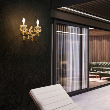 A wall-mounted sconce with two candle-shaped bulbs and crystal accents brightens a textured black wall. Through the glass door, an elegant living room with a leather sofa is visible, while outside, a white lounge chair sits under an Outdoor Crystal Chandelier Wall Light.