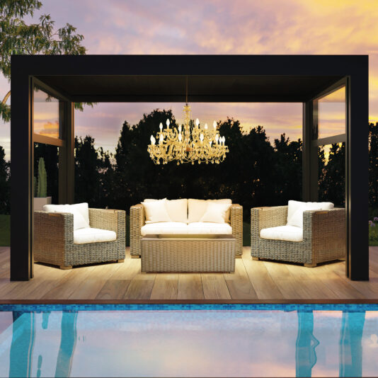 An outdoor seating area features wicker furniture—a sofa and two armchairs—beneath a rectangular pergola. Overhead, an Outdoor Crystal Glass Chandelier elegantly illuminates the scene. The space overlooks a pool, framed by trees and a vibrant sunset sky.