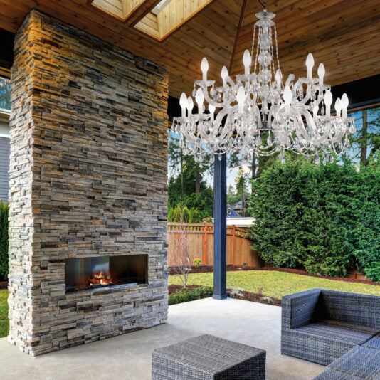 An outdoor patio showcases a stone fireplace beside wicker furniture, highlighted by the Outdoor Crystal Glass Chandelier hanging from a wooden ceiling. The scene is complemented by lush greenery and a quaint wooden fence in the background.
