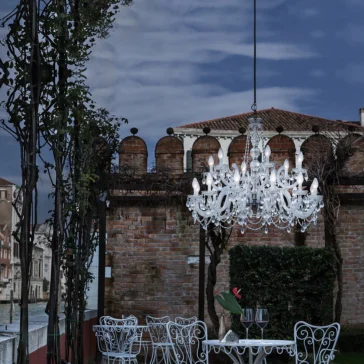 An elegant outdoor dining scene showcases a white metal table and chairs with a wine glass, all under an Outdoor Crystal Glass Chandelier. It's framed by a brick building with arches and a partly cloudy sky.