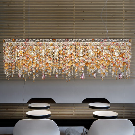 A luxurious Contemporary Rectangular Multicoloured Crystal Chandelier is elegantly suspended above a modern dining table set with plates, while horizontal wooden slats in the background enhance the chic ambience.