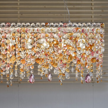 The Contemporary Rectangular Multicoloured Crystal Chandelier, adorned with vibrant crystal beads in shades of orange, pink, and clear, hangs elegantly from the ceiling, casting a shimmering display against a wooden slat background.
