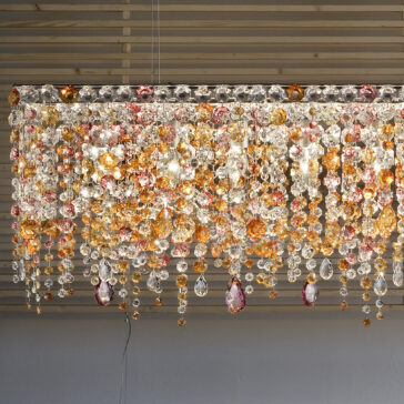 A Contemporary Rectangular Multicoloured Crystal Chandelier featuring cascading crystals in orange, pink, and clear shades is suspended from a slatted ceiling. The illuminated crystals emit a warm and elegant glow, making it an ideal addition to any modern space.