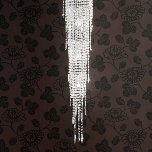 The Long Tiered Crystal Italian Chandelier hangs against a dark, black floral-patterned wall, capturing and reflecting light to produce sparkling highlights.