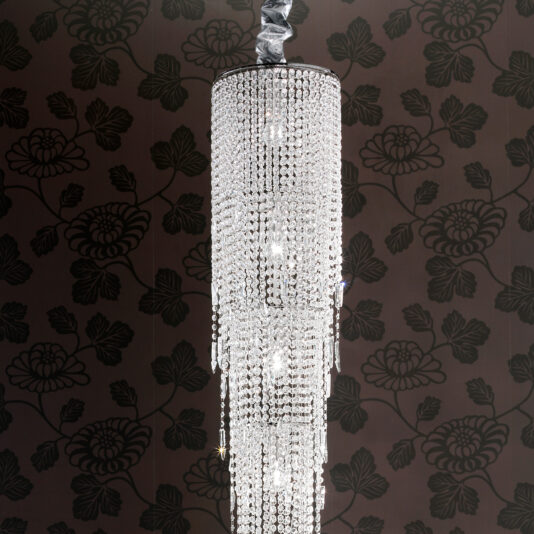 The Long Tiered Crystal Italian Chandelier, with cascading crystal beads, hangs from the ceiling and glows against a dark, floral-patterned wallpaper background.
