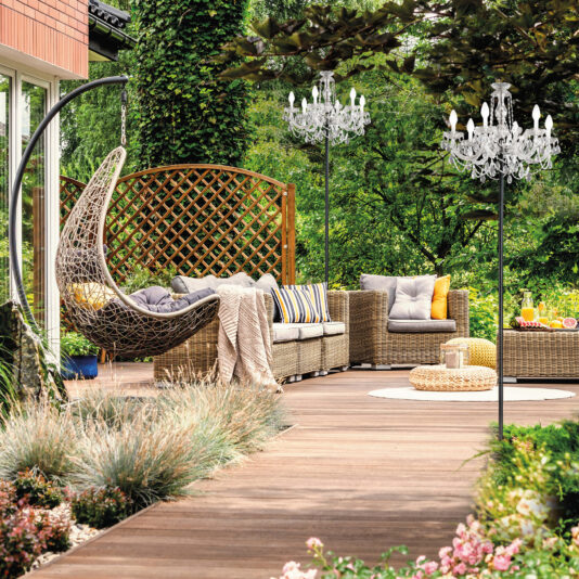 Outdoor Chandelier Floor Lamp A cozy outdoor patio features wicker furniture with colorful cushions beside a lush garden. A swinging egg chair hangs near a textured fence. An Outdoor Chandelier Floor Lamp elegantly lights the area, while a wooden path weaves through the greenery.