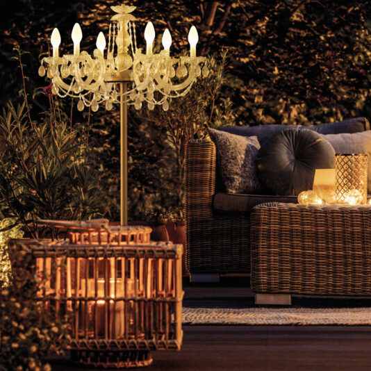 A cozy night-time outdoor seating area features wicker furniture with cushions. An Outdoor Crystal Chandelier Table Lamp hangs above, casting a gentle glow, while decorative candleholders add warmth. Surrounding plants create a serene ambiance.