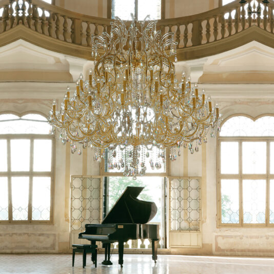 An Extremely Large Classic Gold Crystal Chandelier hangs in an elegant room with a grand piano and matching stool. Tall, arched windows and classical architectural details enhance the luxurious ambiance.