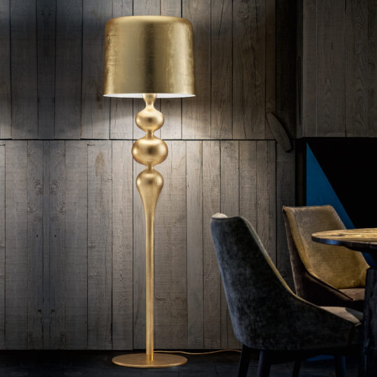 A Designer Modern Gold Leaf Floor Lamp with a sculptural base illuminates a dim room with wooden walls, while nearby dark upholstered chairs and a table enhance the elegant and cozy ambiance.