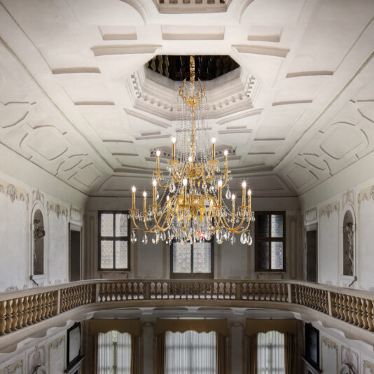 Ornate interior of a grand hall with a Large Italian Gold Plated Crystal Chandelier hanging from the ceiling. The walls feature elaborate moldings, and windows line the upper level. A balcony with a wooden balustrade overlooks the lower floor, which has draped windows.