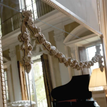 The Luxurious Large Louis Philippe Mirror, adorned with floral designs, reflects a grand room featuring a black piano and a lit lamp. Large windows and architectural details enhance the luxurious atmosphere, adding elegance and refinement to the space.