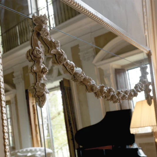 The Luxurious Large Louis Philippe Mirror, adorned with floral designs, reflects a grand room featuring a black piano and a lit lamp. Large windows and architectural details enhance the luxurious atmosphere, adding elegance and refinement to the space.