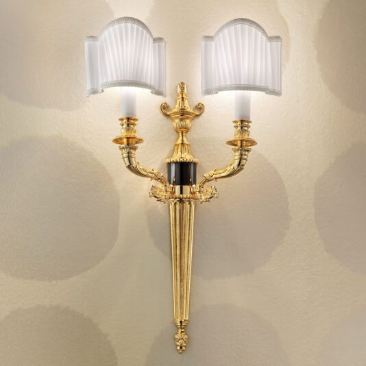 Introducing the Classic Gold Plated Double Wall Light—a vintage-style sconce adorned with ornate details. This elegant fixture features two candle-like arms, each crowned with pleated white lampshades. It is perfectly complemented by a textured wall background with subtle circular patterns.