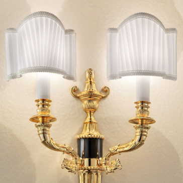 The Classic Gold Plated Double Wall Light is an ornate fixture with two elegantly curved arms, each adorned with a white pleated lampshade. Intricate detailing and a central decorative finial enhance its classic appeal, creating a soft glow against a beige, textured background.