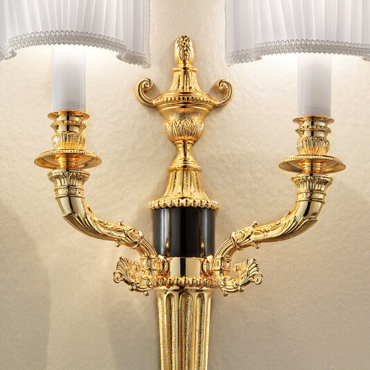 A close-up of the Classic Gold Plated Double Wall Light reveals its ornate, classic design with intricate detailing. This light features two candle-like branches topped with fabric lampshades and a central decorative urn. Mounted on a beige wall, the wall sconce casts a warm glow as the lamps are lit.