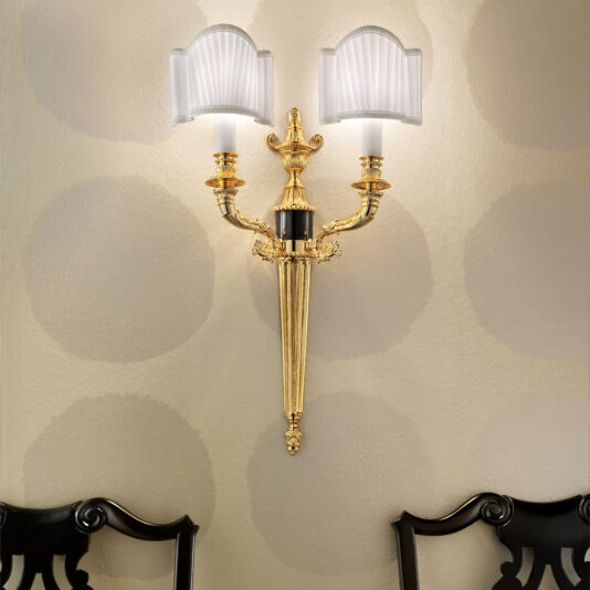 A Classic Gold Plated Double Wall Light with three ornate, gold-plated arms elegantly displays two sophisticated lampshades. It is mounted on a textured beige wall adorned with large, soft circular patterns. The scene is partially framed by two dark-colored wooden chairs featuring intricate back designs at the bottom of the image.