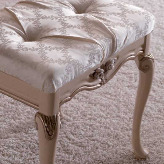 Luxurious Italian Dressing Table Stool A close-up of the Luxurious Italian Dressing Table Stool, showcasing its ornate, cream-colored upholstery with intricate floral patterns on the cushion. The stool features carved details and elegant, curved legs, beautifully placed on a soft, light-colored carpeted floor.
