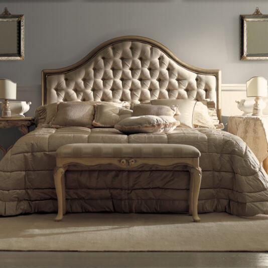 A luxurious bedroom featuring the Classic Bronze Designer Button Upholstered Bed with an ornate, tufted headboard. This designer masterpiece is adorned with multiple pillows and a plush comforter. A matching cushioned bench sits at the foot of the button upholstered bed. Nightstands with lamps flank the bed, and two framed mirrors hang on the wall.