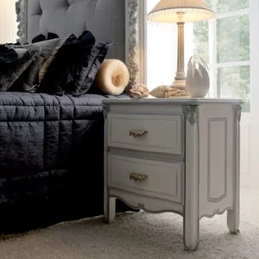 Classic Italian Bedside Table With Drawers A vintage-style bedroom with a Classic Italian Bedside Table With Drawers featuring two ornate handles. The nightstand holds a decorative lamp with a pleated shade and small sculptures. The bed has a tufted headboard, dark blue bedding, and plush pillows in various shades.