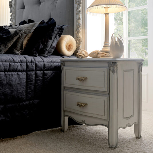 A vintage-style bedroom with a Classic Italian Bedside Table With Drawers featuring two ornate handles. The nightstand holds a decorative lamp with a pleated shade and small sculptures. The bed has a tufted headboard, dark blue bedding, and plush pillows in various shades.