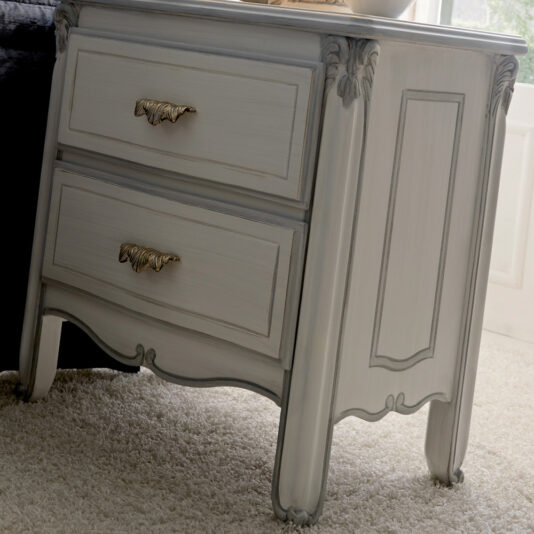 A Classic Italian Bedside Table With Drawers in light gray features two drawers adorned with ornate gold handles. The nightstand boasts carved detailing on the sides and bottom edges, and it is displayed on a light beige carpet in a naturally lit room.