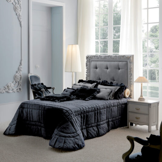 A chic bedroom featuring a decorative tufted headboard, dark bedding, and ruffled pillows. A tall floor lamp stands beside a classic chair in the corner. The Classic Italian Bedside Table With Drawers sits next to the bed, adorned with a small lamp. Large windows with translucent curtains let in natural light.
