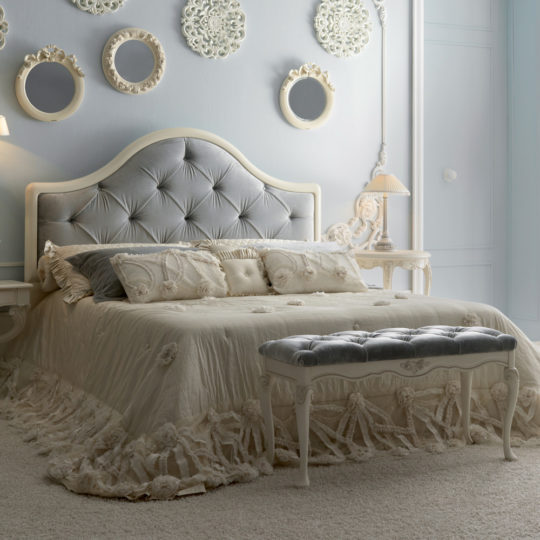 A luxurious bedroom featuring the Classic Italian Designer Button Upholstered Bed with an elegant light gray tufted headboard, complemented by cream-colored bedding adorned with intricate lace details and a matching tufted bench at the foot. The walls are embellished with decorative round mirrors and ornate frames, creating a sophisticated ambiance.
