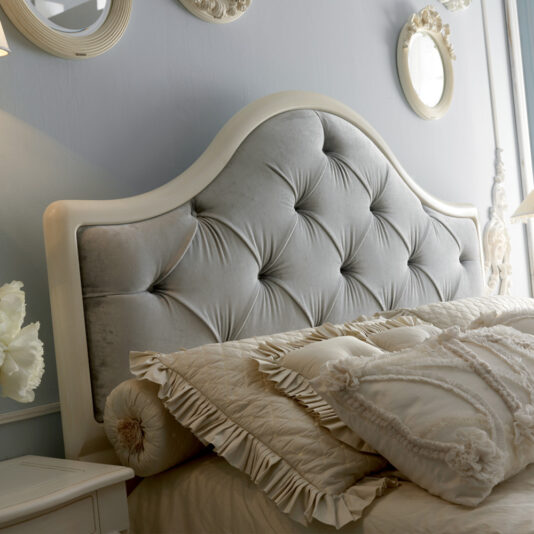 A close-up of a luxurious bedroom featuring the Classic Italian Designer Button Upholstered Bed with an elegant tufted headboard in soft, smooth fabric. The bedding includes various decorative pillows and a quilted blanket in shades of cream. The wall is adorned with several ornate framed mirrors and artwork.