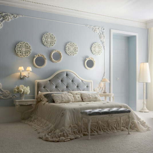 An elegant bedroom with light blue walls adorned with decorative white patterns and round framed mirrors. The room features a Classic Italian Designer Button Upholstered Bed with plush bedding, a matching bench, wall sconces, bedside tables, a floor lamp, and ornate detailing throughout.