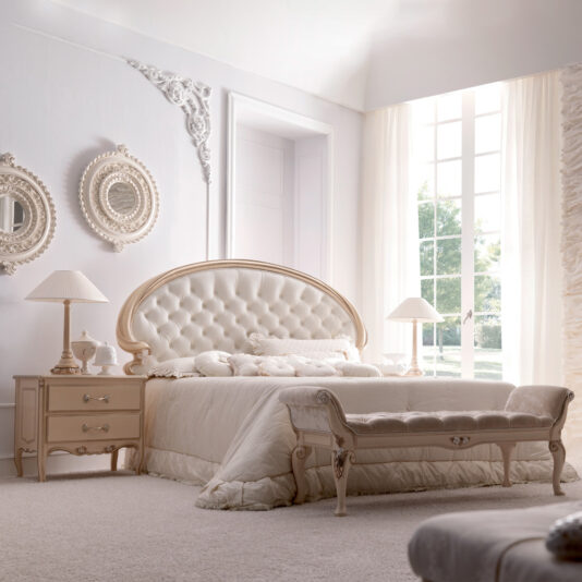 Classic Italian Rose Design Vermeil Bedside Table A beautifully decorated bedroom featuring a large tufted headboard bed with light bedding. The room includes two Classic Italian Rose Design Vermeil Bedside Tables with lamps, a bench at the foot of the bed, ornate mirrors on the wall, and a large window with sheer curtains letting in natural light.