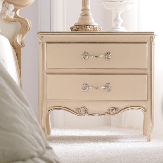 Classic Italian Rose Design Vermeil Bedside Table The Classic Italian Rose Design Vermeil Bedside Table in a vintage cream color, adorned with ornate handles, stands in front of light-colored curtains. On top of the bedside table is a matching decorative lamp and an elegant glass dish. A plush bed partially appears on the left side.