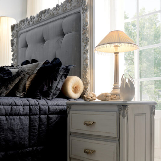 A cozy bedroom scene showcasing the Classic Reproduction Button Upholstered Bed, complete with a tufted gray headboard and intricate detailing. The bed is elegantly made with dark gray bedding and numerous decorative pillows. Adjacent to the bed is a chic bedside table adorned with a vintage lamp and various decorative items, perfectly complementing this timeless design.