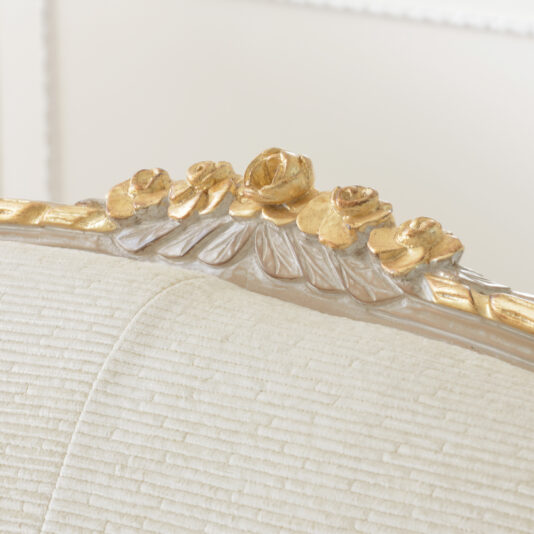 Cream And Gold Reproduction Louis Armchair Close-up of the top part of the Cream And Gold Reproduction Louis Armchair. The frame features a decorative carving of golden roses and leaves, smoothly integrated into the light-colored wood. The chair is textured with horizontal lines, adding contrast to the ornate frame.