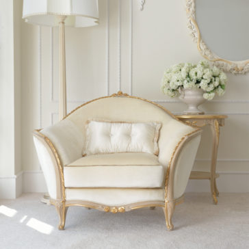 Cream And Gold Reproduction Louis Armchair A Cream And Gold Reproduction Louis Armchair rests on a light carpet, paired with a matching cushion. Next to it, a round side table supports a glass vase filled with white flowers. An elegant tall floor lamp and an ornate wall mirror complete the sophisticated setting.