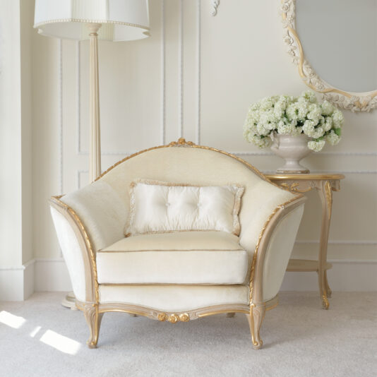 Cream And Gold Reproduction Louis Armchair A Cream And Gold Reproduction Louis Armchair rests on a light carpet, paired with a matching cushion. Next to it, a round side table supports a glass vase filled with white flowers. An elegant tall floor lamp and an ornate wall mirror complete the sophisticated setting.