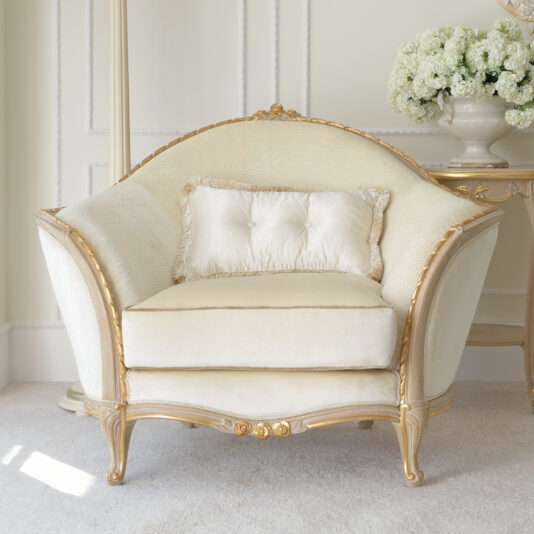 A luxurious Cream And Gold Reproduction Louis Armchair with ornate detailing stands in an elegantly decorated room. A matching pillow rests on the chair. Behind it, a side table with a large vase of white flowers completes the sophisticated setting.