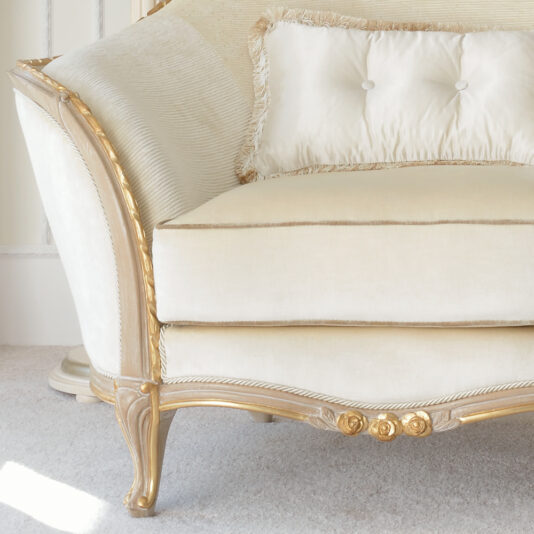 Cream And Gold Reproduction Louis Armchair An elegant white upholstered sofa with beige and gold accents features a tufted back cushion with button details and fringe trim. Reminiscent of the Cream And Gold Reproduction Louis Armchair, the sofa boasts ornately carved wooden legs and armrests adorned with intricate floral motifs at the base. It is placed on a light carpet.