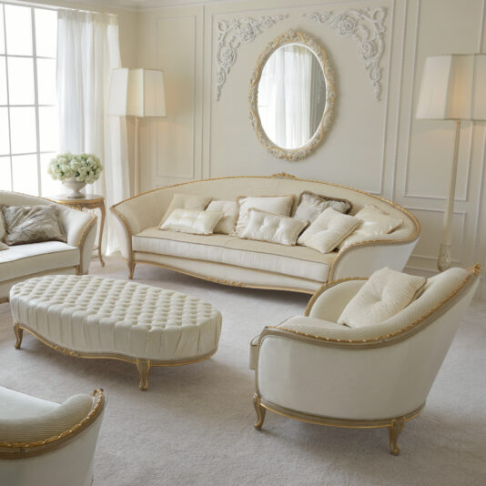 Cream And Gold Reproduction Louis Armchair An elegant living room with classic cream and gold furniture. It features a sofa, two Cream And Gold Reproduction Louis Armchairs, an upholstered coffee table, standing lamps, and an ornate wall mirror. The room is bathed in natural light from a large window with sheer curtains.