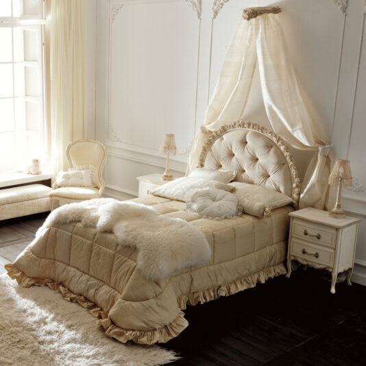 A luxurious bedroom showcases an ornate cream-colored Floral Ivory And Gold Single Italian Bed, boasting a tufted headboard and an elegant canopy. The bed is adorned with decorative pillows and a fur throw, flanked by two nightstands with lamps. Nearby, a cushioned chair and a window bench complete the sophisticated setting.