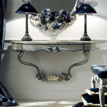 French Rose Wall Fixing Console Table An elegant French Rose Wall Fixing Console Table, featuring a curved, carved design in an antique finish, is mounted on a light-colored wall. Adorning the table are two black-shaded lamps and a floral arrangement positioned at the center. Above it hangs a rounded mirror, while to the right, a partially visible cushioned stool can be seen.