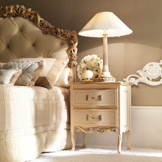 High End Italian Rose and Ribbon Button Upholstered Bed A luxurious bedroom features the High End Italian Rose and Ribbon Button Upholstered Bed, showcasing an ornate beige headboard with intricate floral carvings. A matching bedside table with two drawers, similarly adorned with elaborate carvings, supports a decorative lamp with a pleated shade and a vase of white roses. The atmosphere is elegant and serene.