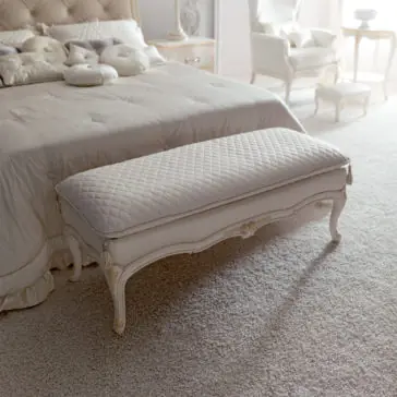 Italian Designer Bedroom Bench A serene bedroom featuring a tufted headboard bed adorned with plush pillows and a pastel bedspread. At the foot of the bed stands an **Italian Designer Bedroom Bench**, elegant and upholstered. The room is bathed in soft natural light, highlighting the pastel decor and ornate furniture.