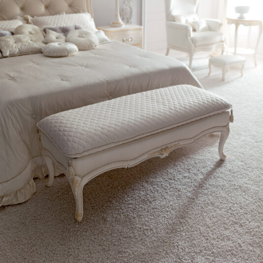 A serene bedroom featuring a tufted headboard bed adorned with plush pillows and a pastel bedspread. At the foot of the bed stands an **Italian Designer Bedroom Bench**, elegant and upholstered. The room is bathed in soft natural light, highlighting the pastel decor and ornate furniture.