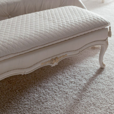 Italian Designer Bedroom Bench A close-up view of the Italian Designer Bedroom Bench reveals its elegant quilted white fabric upholstery, complemented by ornate wooden legs with intricate carvings. The bench stands gracefully on a plush, light-colored carpet.