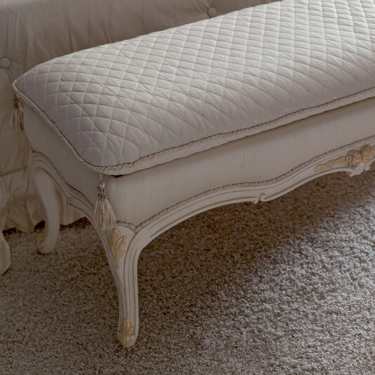 Italian Designer Bedroom Bench The Italian Designer Bedroom Bench, exuding a vintage elegance with its white quilted upholstery and ornate wooden legs, is detailed with subtle light gold accents. It is placed on a plush, light beige carpet and set against a matching fabric backdrop.