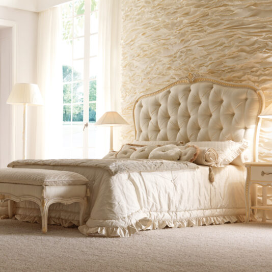 Luxurious bedroom showcasing an elegant Italian Ivory and Gold Luxury Reproduction Bed set against a textured wall. The room features matching bedside tables and lamps, a padded bench at the foot of the bed, and tall windows that flood the space with natural light, creating a serene ambiance.