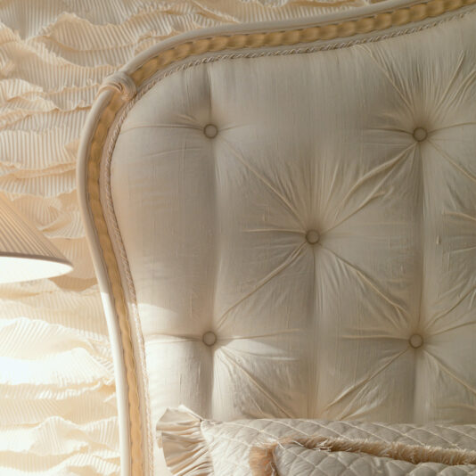 Italian Ivory And Gold Luxury Reproduction Bed Here is a close-up of the Italian Ivory And Gold Luxury Reproduction Bed's elegant, tufted headboard in a luxurious cream-colored upholstery, featuring intricate stitching and button details. The coordinating pillows in matching textured fabric are partially visible in front of the headboard, with a lampshade seen on the left side.